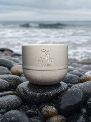 Ocean Mug in Sand – sandfarbene Steinzeug-Tasse auf Steinen vor sanften Wellen